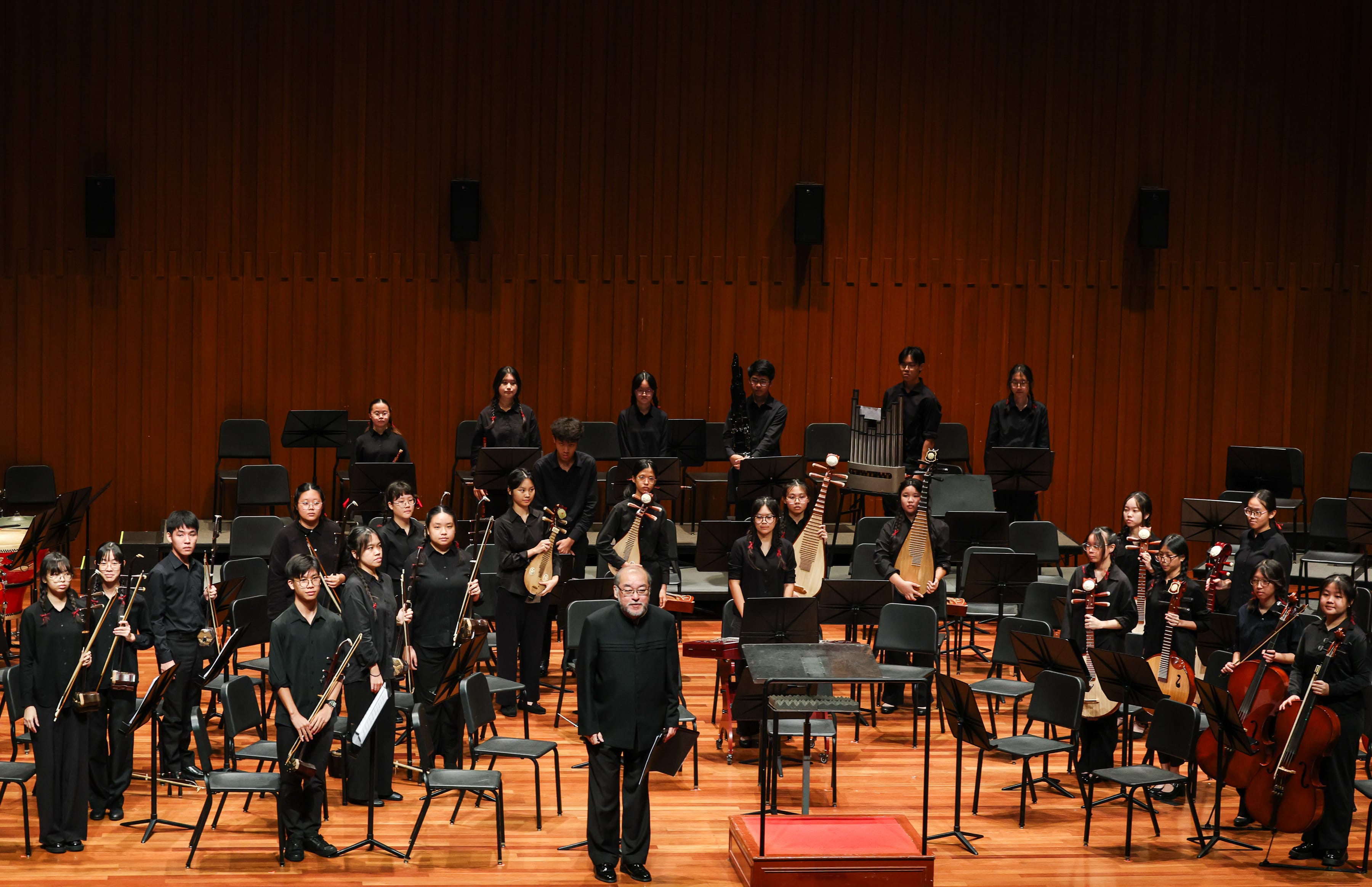 A large student Chinese orchestra stands on a formal concert stage with warm wooden panelling. The students, all dressed in black, are arranged in sections across the stage holding various traditional Chinese instruments. A conductor in black stands at the front centre on a podium, facing the audience.